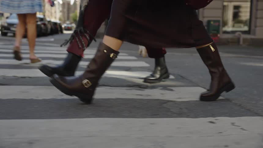 A slow-motion close-up follows the legs of two stylishly dressed people in leather boots as they walk across a sunny city street