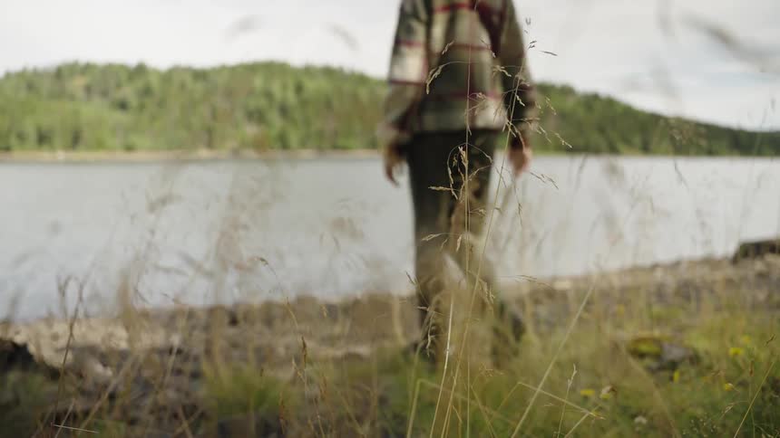 A woman in a plaid jacket and backpack walks along the shore of a serene lake surrounded by a forest