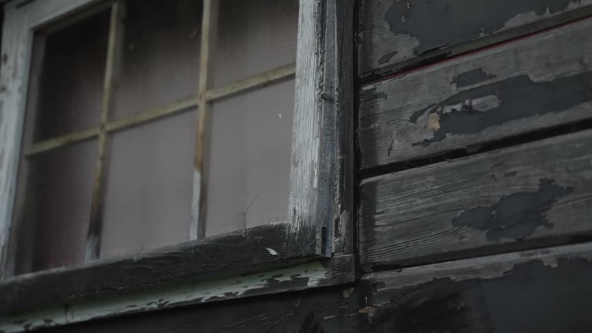 A slow tilt-up shot reveals the peeling paint and weathered frame of an old, eerie window on a wooden house