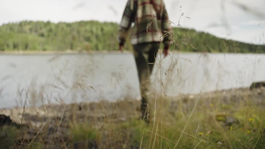 A hiker walks in slow motion towards a tranquil lakeside, with tall grass in the foreground