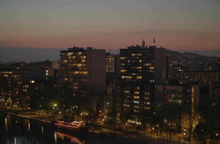 Wide shot of a tranquil Parisian cityscape at dusk, with lights illuminating apartment windows and a tour boat on the canal