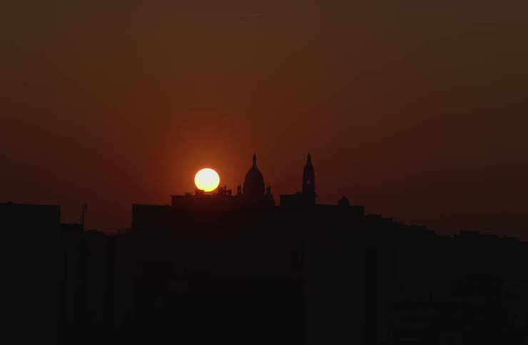 An orange sun sets in a dramatic time-lapse behind the silhouette of a city skyline featuring a domed cathedral