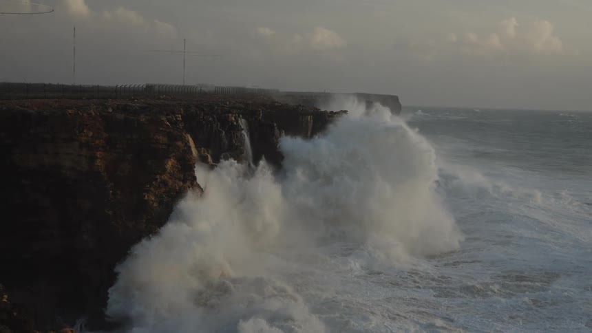 Massive ocean waves crash against a rugged cliffside in dramatic slow motion during a powerful storm