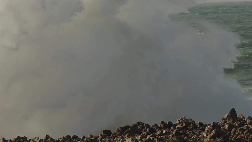 Slow motion footage shows a massive ocean wave crashing against a rocky shoreline, creating a giant wall of white water spray