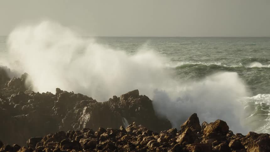 A massive ocean wave crashes against a rocky coastline in dramatic slow motion