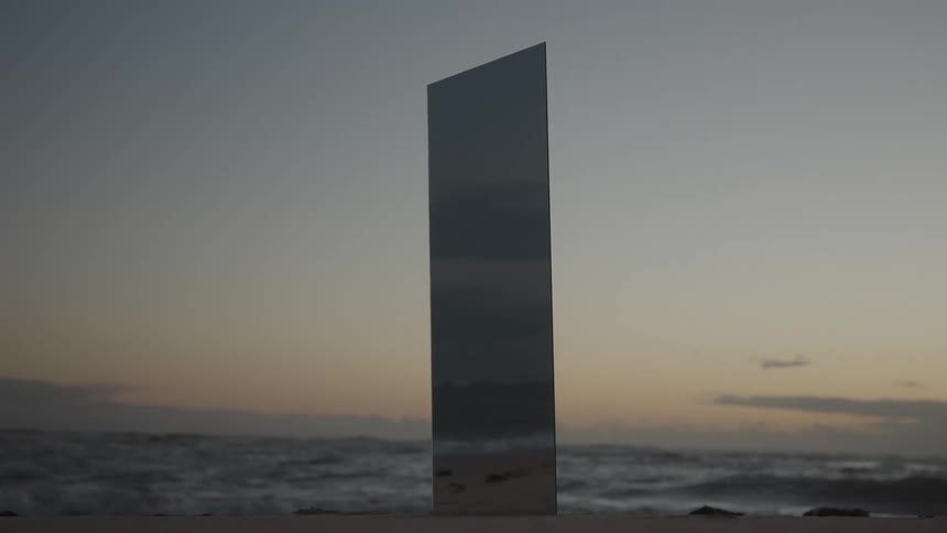 A mysterious mirrored monolith stands on a tranquil beach at dawn with the ocean in the background