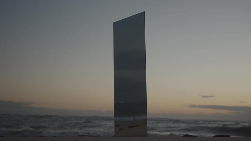 A mysterious mirrored monolith stands on a desolate beach with ocean waves in the background at dusk