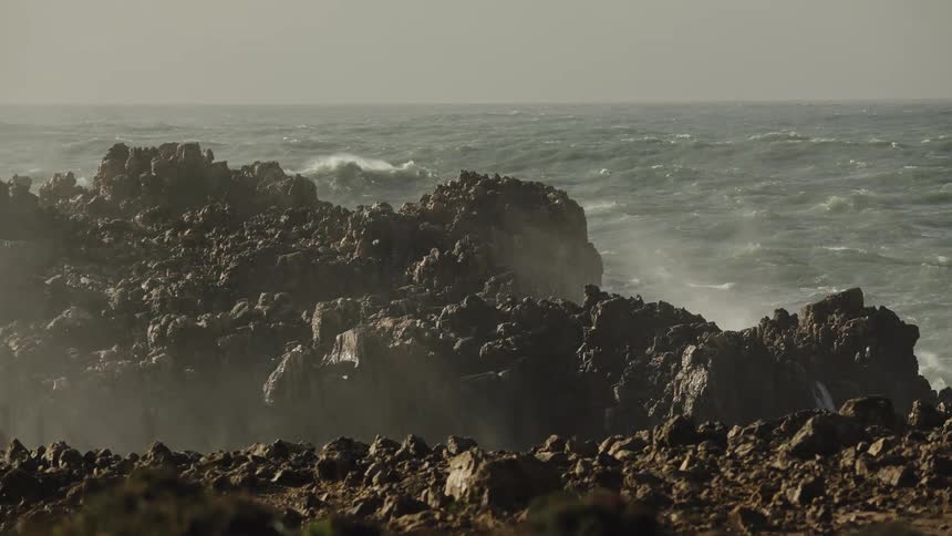 Slow dolly shot of misty sea spray blowing over rugged rocks as the powerful ocean waves crash against the shore