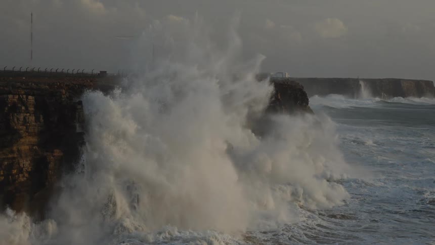 A dramatic slow-motion shot captures the raw power of huge ocean waves crashing violently against a rugged cliffside