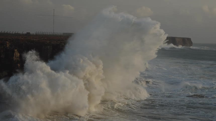 A massive ocean wave crashes against a rugged cliff in dramatic slow motion during a powerful storm at dusk