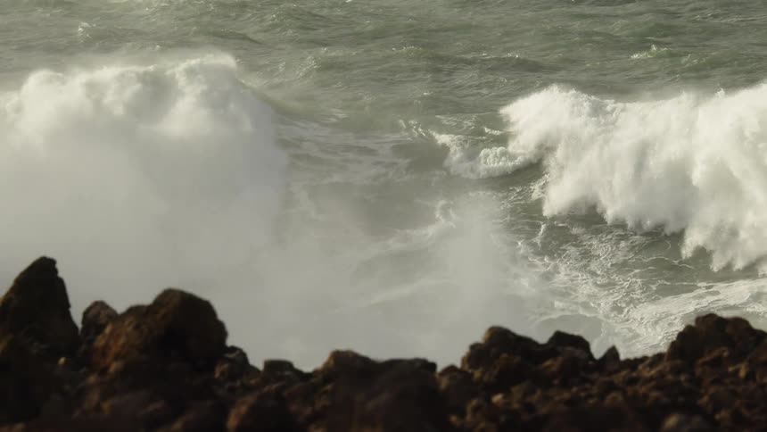 A massive ocean wave crests and crashes in slow motion as seen from a rocky shoreline
