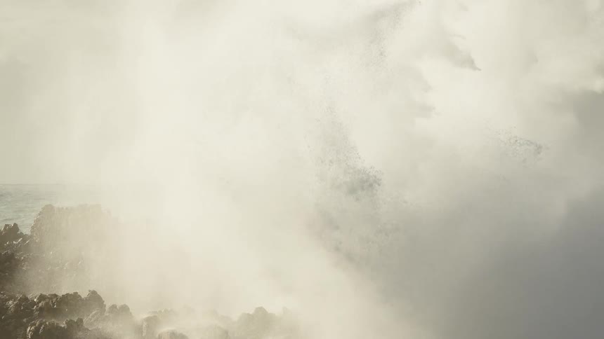A giant ocean wave explodes against a rugged, rocky shoreline in dramatic slow motion