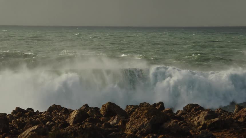 A massive ocean wave crashes against a rocky shoreline in dramatic slow motion during a storm