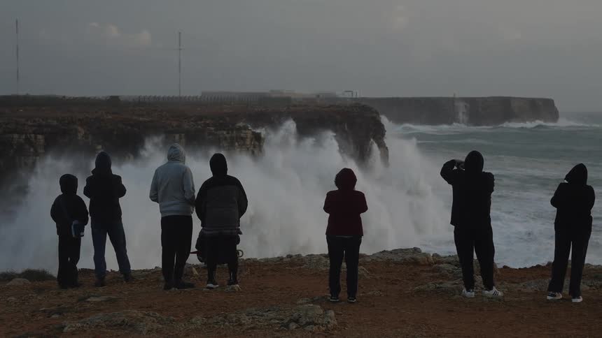 A group of onlookers in hoodies stand on a clifftop, watching and filming as massive storm waves crash against the rocky coastline