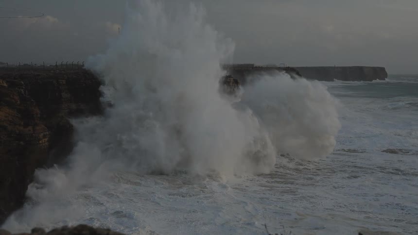 Dramatic slow motion footage shows massive ocean waves crashing against a rocky cliffside during a storm