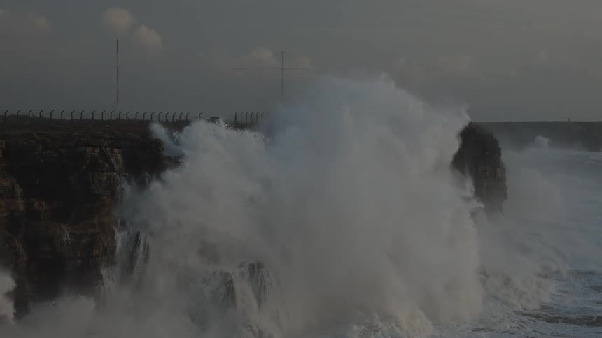 Massive ocean waves crash against a rugged cliffside in dramatic slow motion, creating powerful waterfalls during a storm