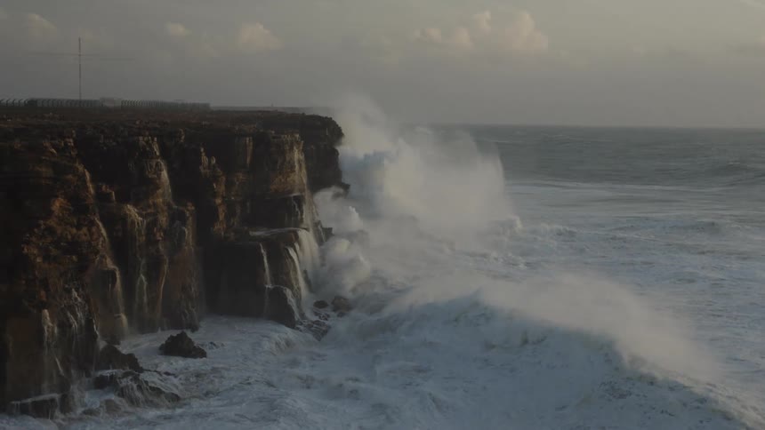 Slow motion shot of massive ocean waves from a storm crashing against a rocky cliff side during an overcast day