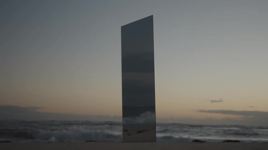 A mysterious mirrored monolith stands on a sandy beach as ocean waves roll in at sunrise