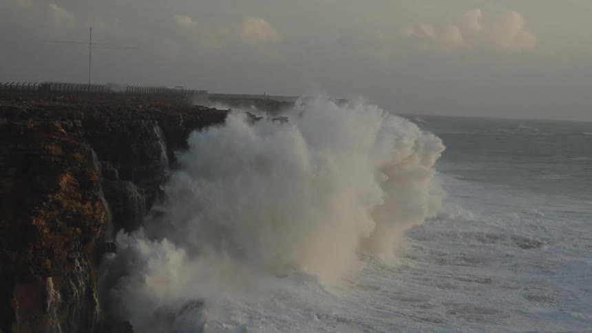 Dramatic slow-motion footage shows massive, powerful ocean waves crashing against a rugged cliffside during a storm