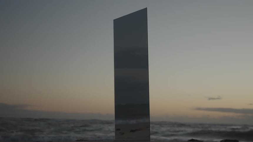 A mysterious mirrored monolith stands on a sandy beach as ocean waves roll in during a tranquil sunset