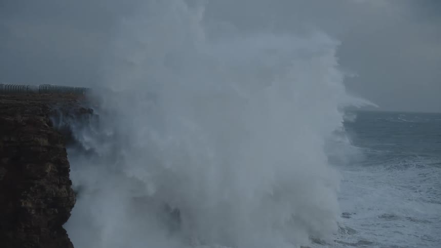 A massive ocean wave crashes against a rugged cliff face in dramatic slow motion, creating a spectacular explosion of whitewater