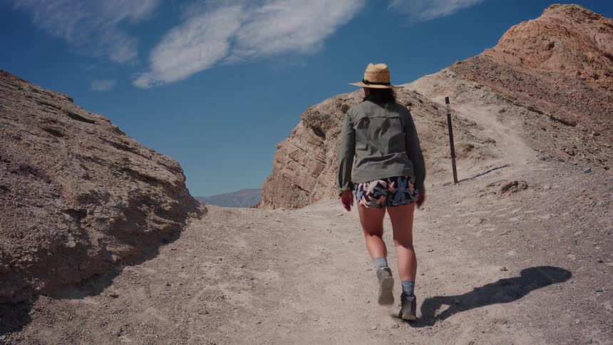A woman in a straw hat walks away from the camera in slow motion up a dusty trail in a rocky desert canyon