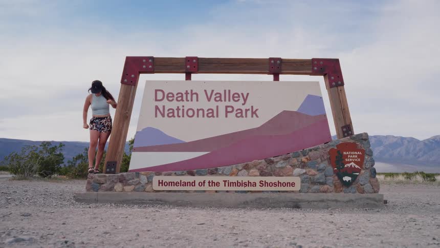 A woman playfully poses for pictures next to the iconic Death Valley National Park sign in the desert