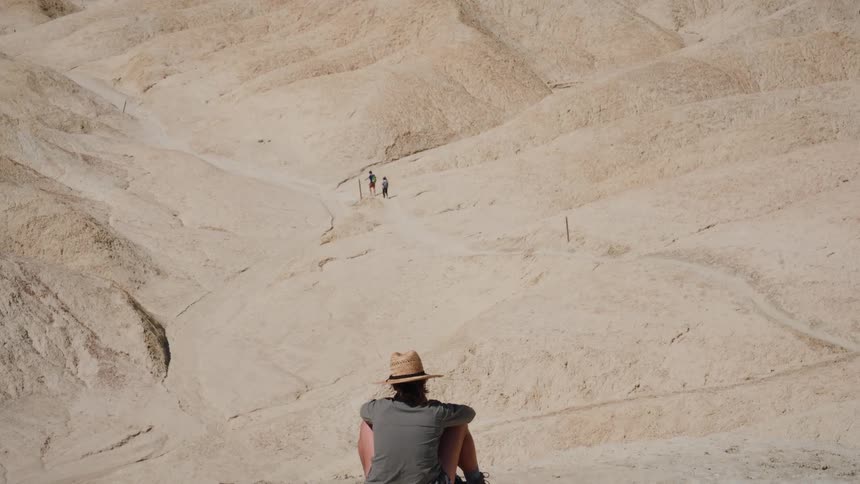 A lone hiker with a hat sits on a sandy overlook, watching other people explore the vast, sun-drenched badlands of a desert canyon
