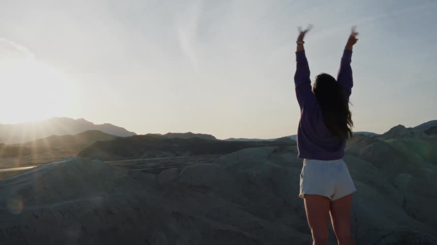 A young woman stands on a mountain peak at sunset, stretching her arms towards the sky in slow motion
