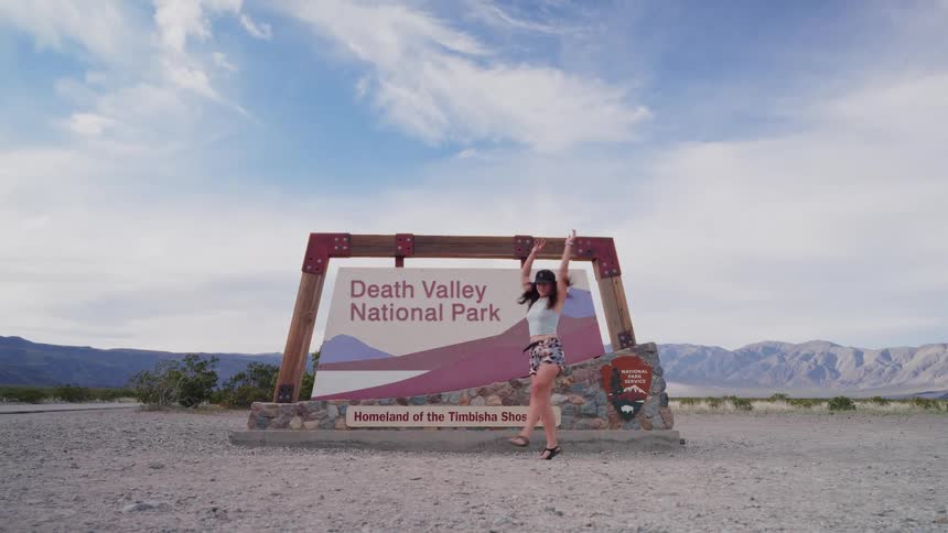A joyful woman dances playfully in front of the Death Valley National Park entrance sign under a wide desert sky