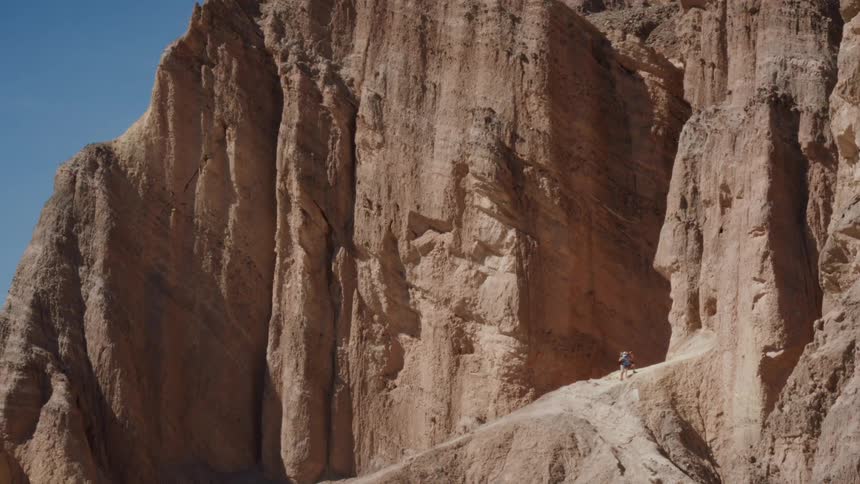 A fast zoom reveals a lone hiker on a narrow trail, dwarfed by the immense rock formations of a desert canyon