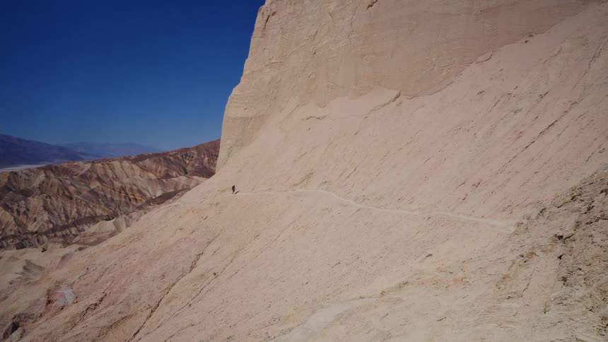 A lone hiker makes their way along a narrow trail etched into the side of a massive desert mountain in Death Valley National Park