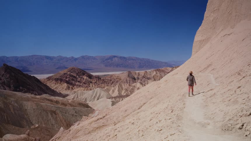 A woman wearing a hat hikes in slow motion along a narrow trail through the colorful desert badlands of Death Valley National Park