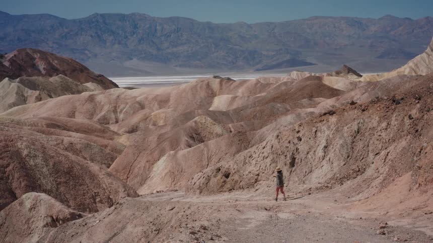 A woman in a hat walks along a trail through the colorful, arid badlands of a vast desert landscape on a sunny day