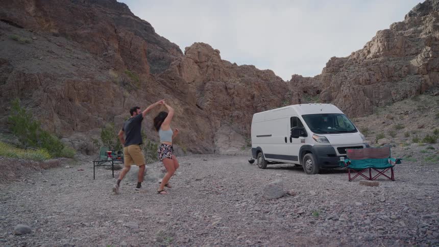 A joyful young couple dances and embraces in slow motion at their remote campsite in a rocky canyon with their camper van