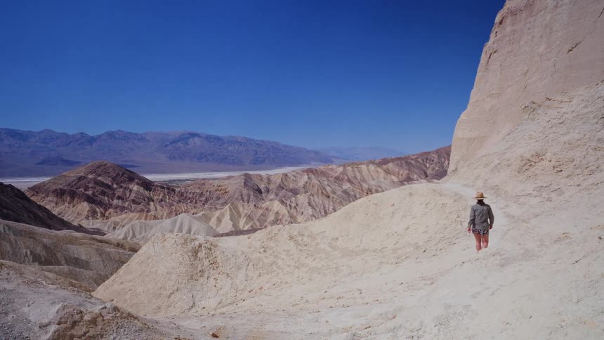 A solo female hiker wearing a straw hat walks in slow motion along a trail through a vast and colorful desert canyon on a sunny day