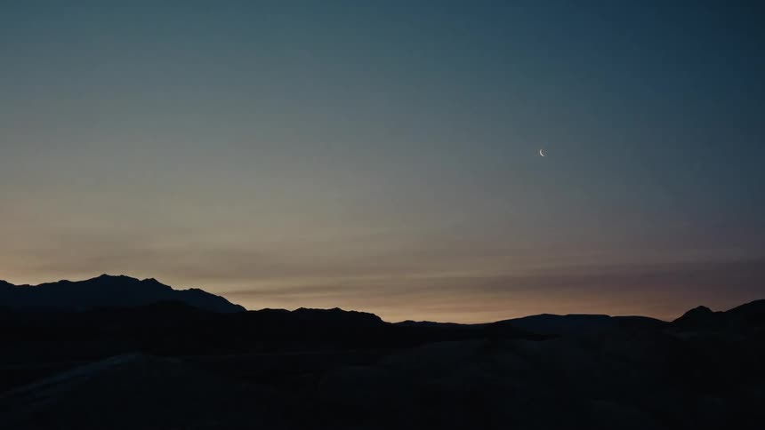 Time-lapse captures the transition from a dark night sky with a crescent moon to a beautiful sunrise over a desert mountain range