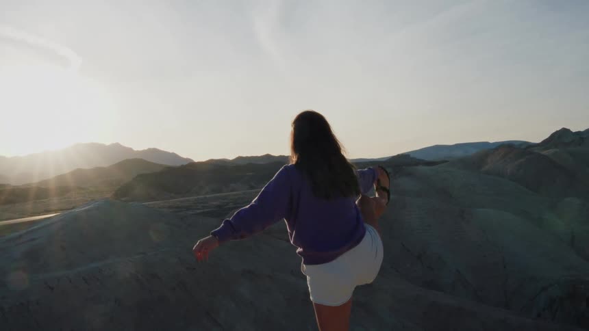 A woman in a purple sweatshirt gracefully finds her balance in a yoga pose while overlooking a vast desert landscape at sunset