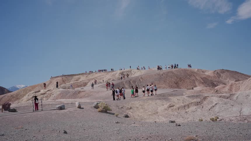 A wide shot shows groups of tourists hiking up a dusty trail on a desert hill at Zabriskie Point in Death Valley National Park