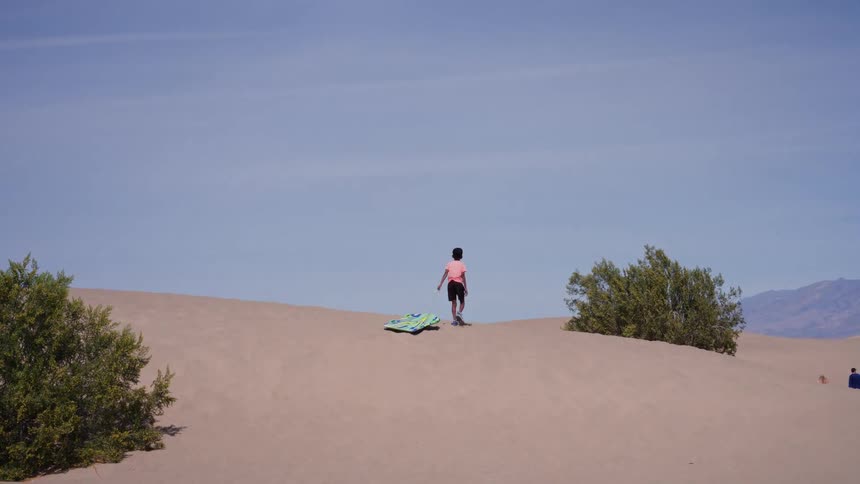A young boy walks up a large desert sand dune in slow motion, pulling a sandboard behind him on a sunny day