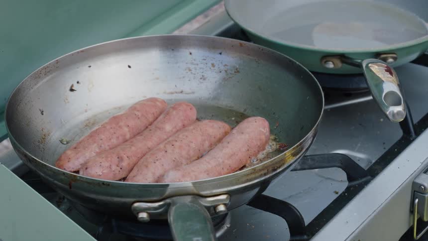 Close-up of four sausages sizzling in hot oil in a metal pan on a portable camping stove