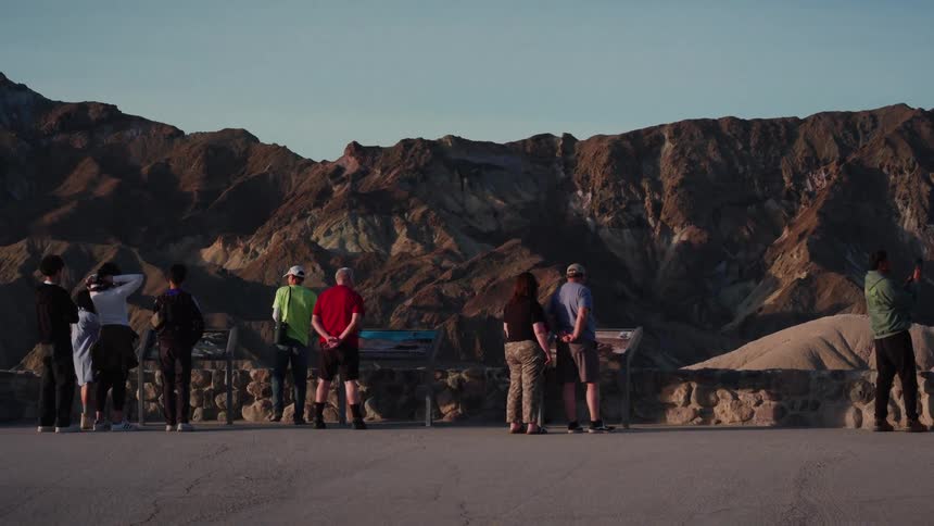 A group of tourists gather at an overlook to admire the colorful, layered mountains of Death Valley National Park at sunrise