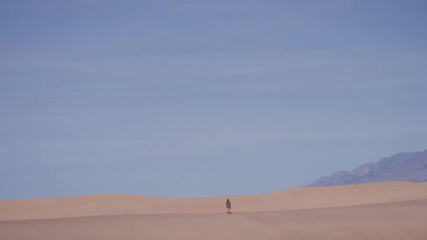 A solitary traveler walks across the expansive sand dunes in the desert with mountains in the distance