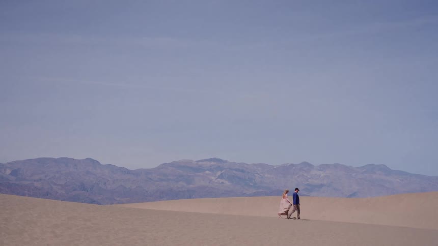 A couple walks across a vast desert sand dune with a picturesque mountain range in the background