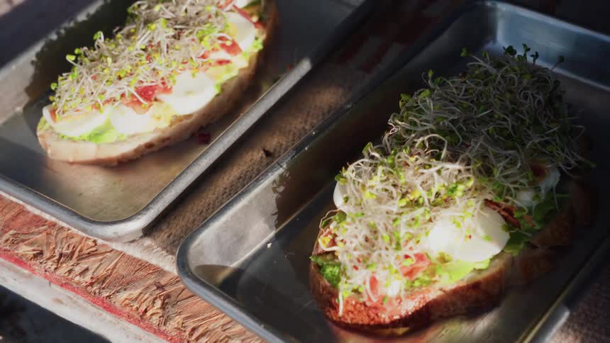 Freshly prepared avocado toast with egg and sprouts sits on metal trays in the morning light