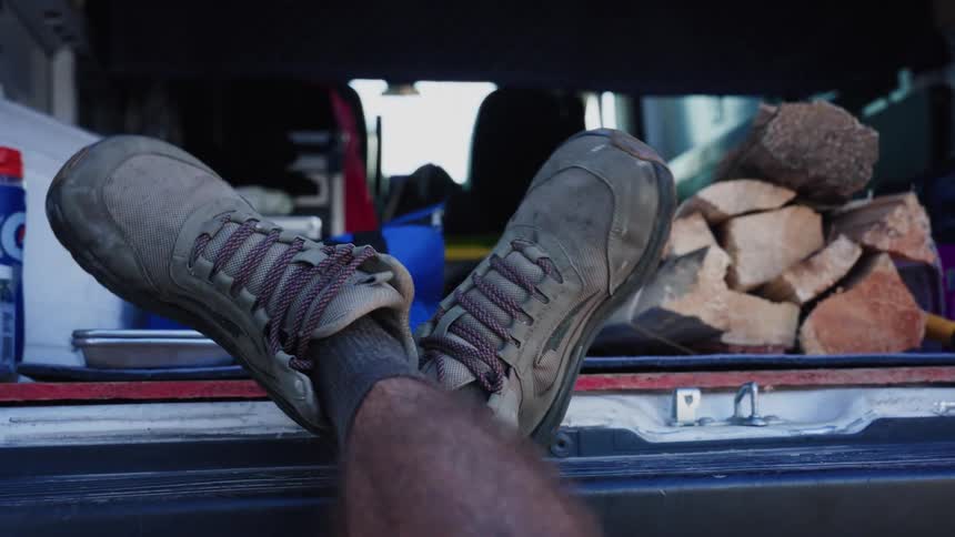A person wearing hiking boots relaxes with their feet up in the back of a van, wiggling their ankles next to a bundle of firewood