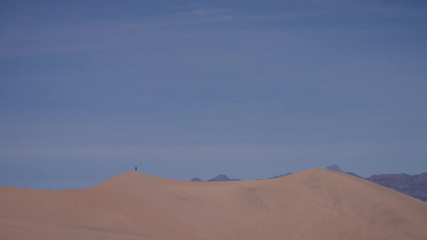 A solitary hiker stands on the crest of a massive sand dune, dwarfed by the immense desert landscape and wide blue sky