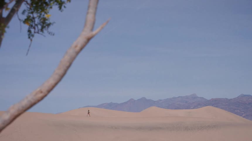 A lone person walks across the vast sand dunes of a desert with distant mountains under a blue sky