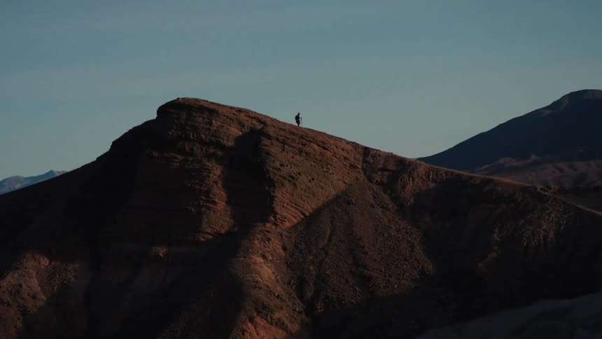 A lone hiker slowly traverses a dramatic mountain ridgeline as the sun casts long shadows across the vast, arid landscape