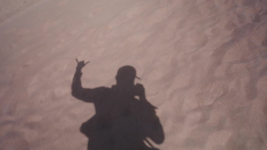 The shadow of a person taking a selfie on the sand makes playful hand gestures like 'hang loose' and 'rock on' signs